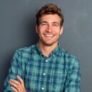 Young handsome man leaning against grey wall with arms crossed. Cheerful man laughing and looking at camera with a big grin. Portrait of a happy young man standing over grey background.